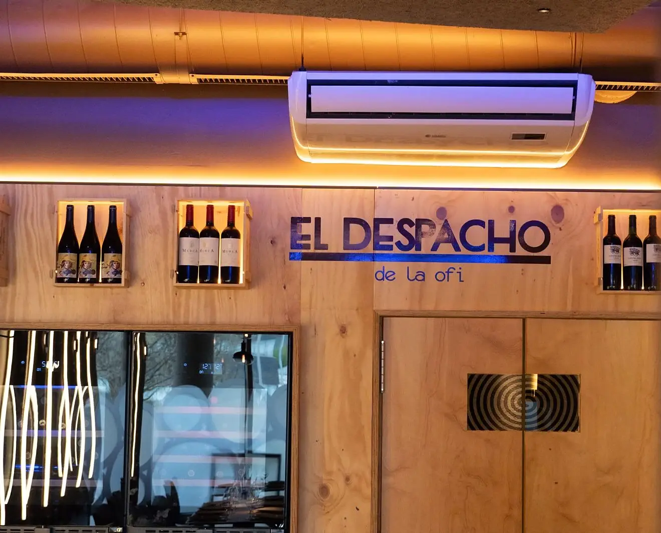 Pared de madera de El Despacho con el rótulo «El Despacho de la ofi», cajas de vino iluminadas y la vinoteca